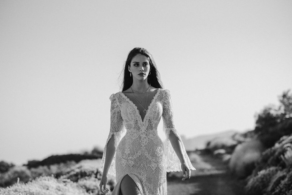 Elegant bride in long-sleeve lace wedding gown with deep V-neckline walking outdoors in black and white photo.