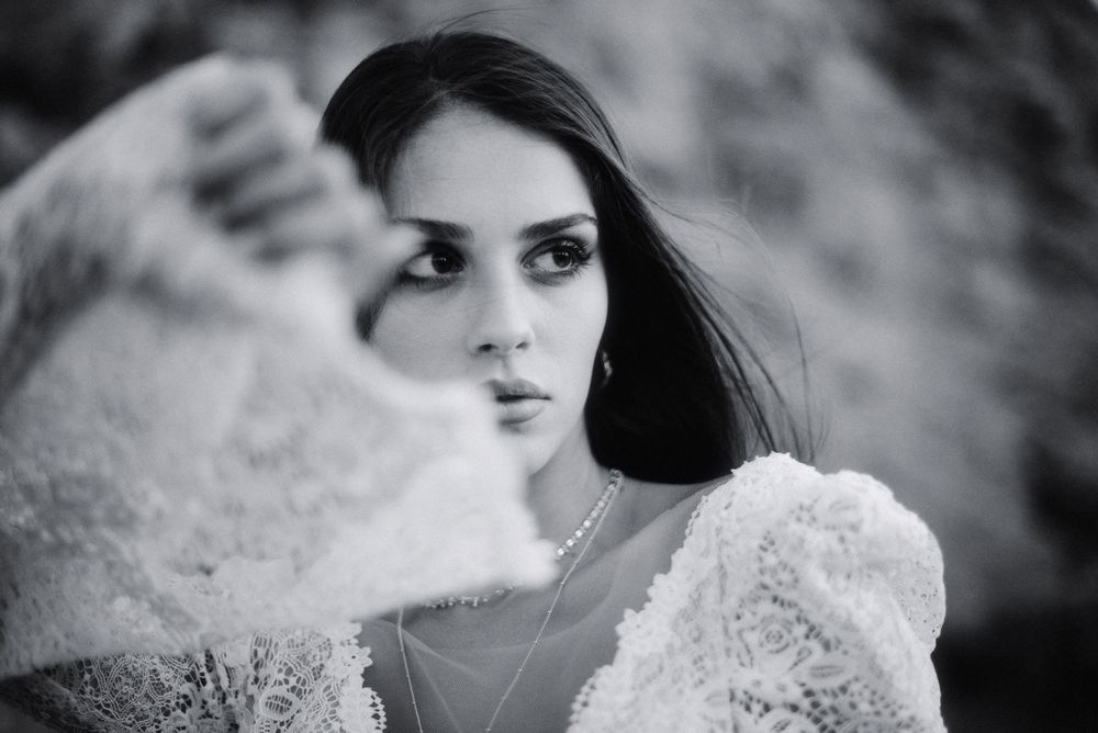 Black and white portrait of a brunette woman in lace dress holding fabric toward camera with a soft bokeh background.