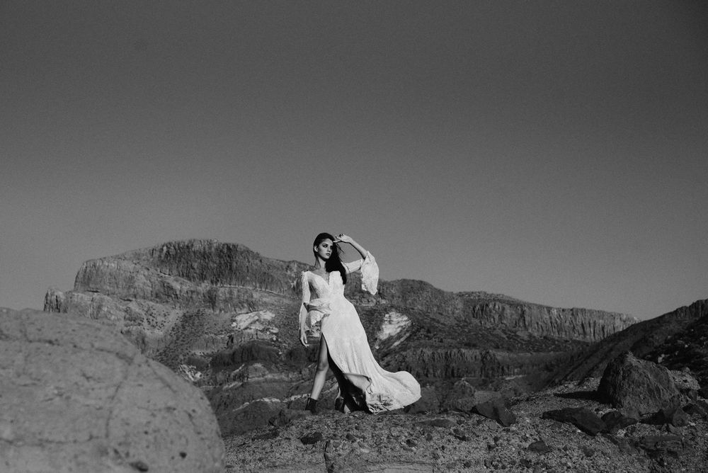 Woman in flowing white gown poses dramatically on rocky desert landscape with mountain backdrop in black and white.