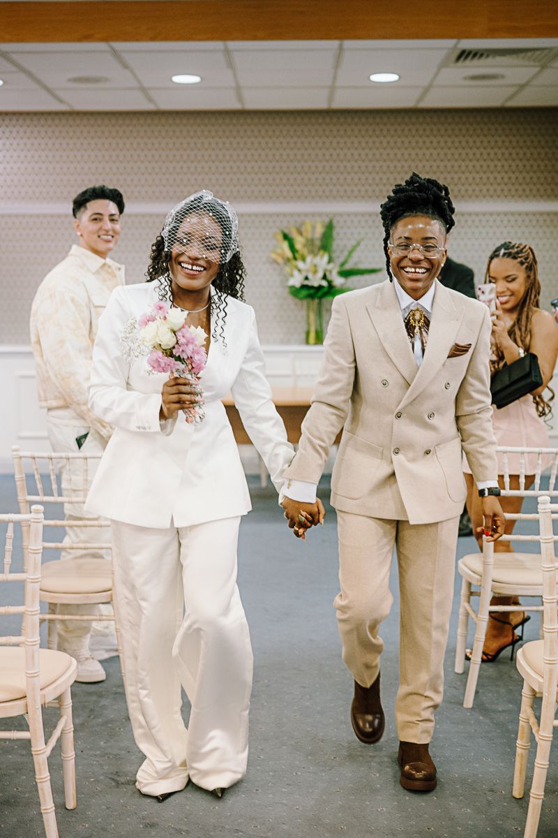 Two brides in white and beige suits walk hand-in-hand down the aisle after their wedding ceremony.