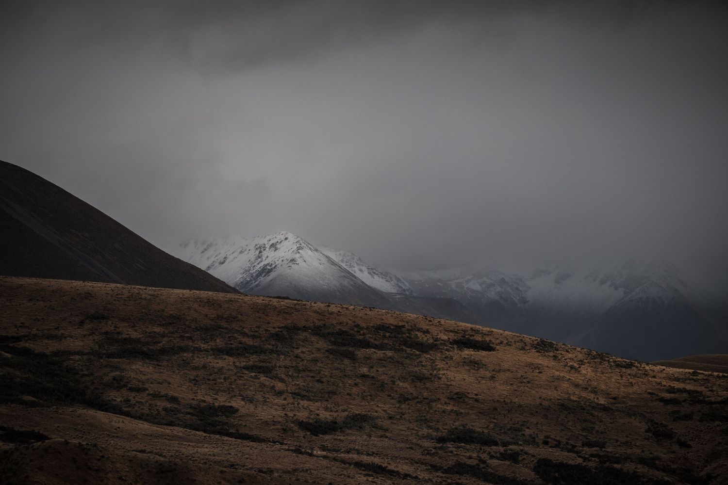 Snow-capped mountains under dark storm clouds with rugged brown hillside in dramatic moody landscape.