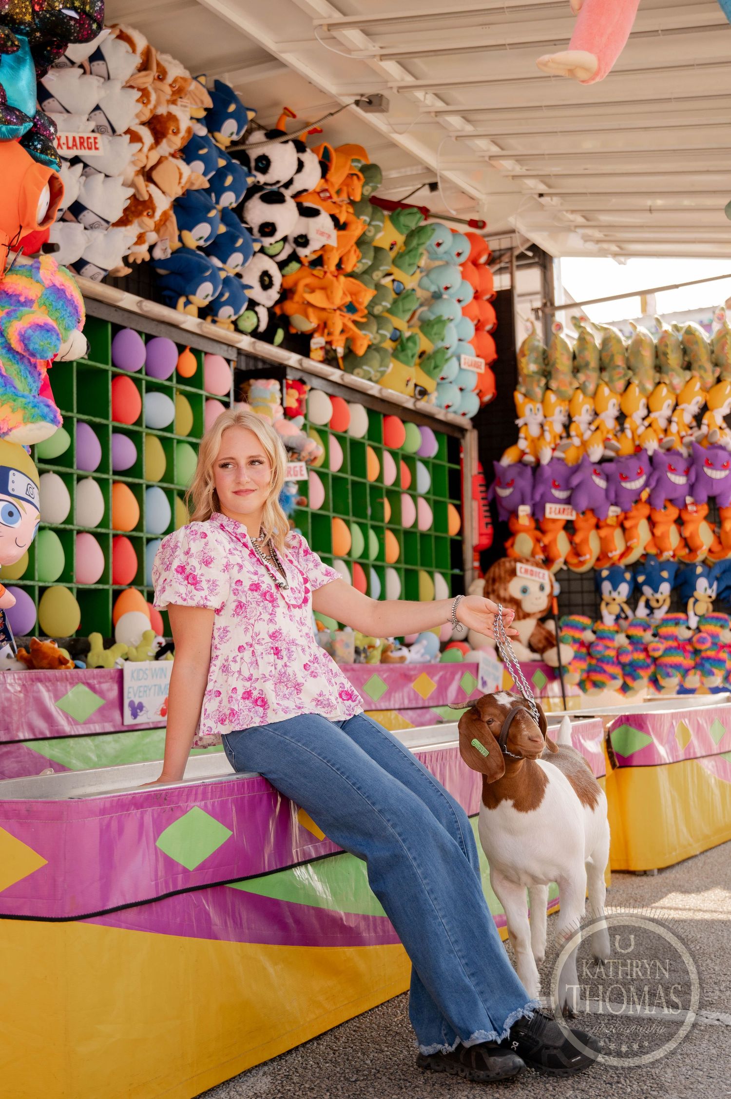 Woman in floral top sits with a goat at a colorful carnival game booth filled with stuffed animal prizes.