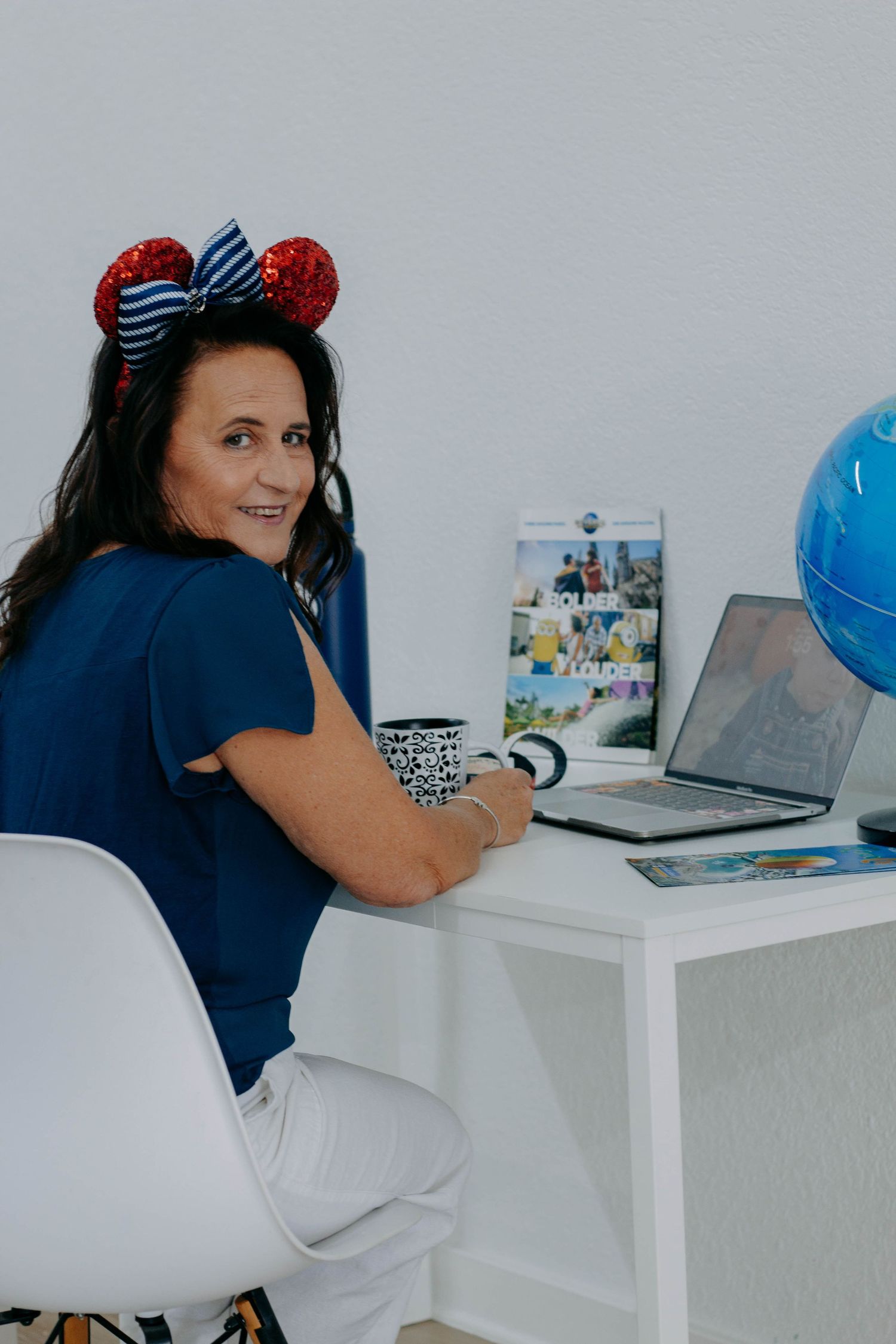 Woman in Mickey Mouse ears sits at desk with laptop, globe, and travel photos, smiling over shoulder.