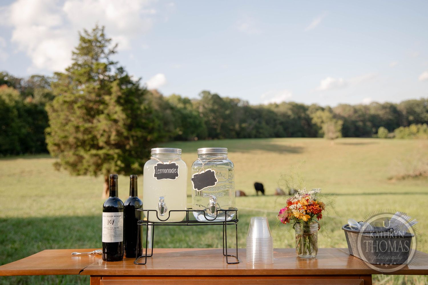 Rustic outdoor wedding drink station with mason jar dispensers, wine bottle, and floral arrangement on wooden table in green field.
