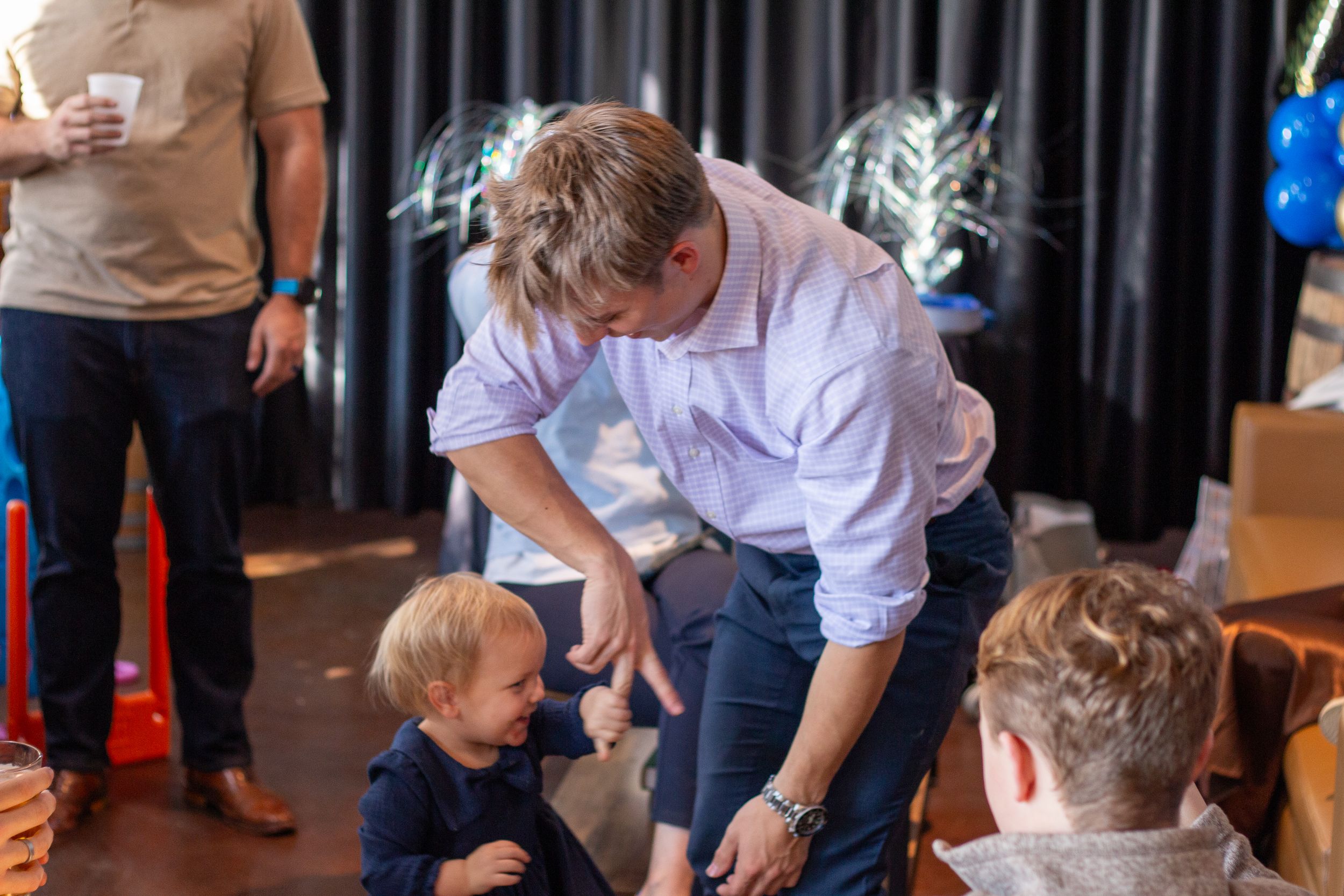 A man in a white shirt bends down to interact with young children at an indoor event with dark curtains.
