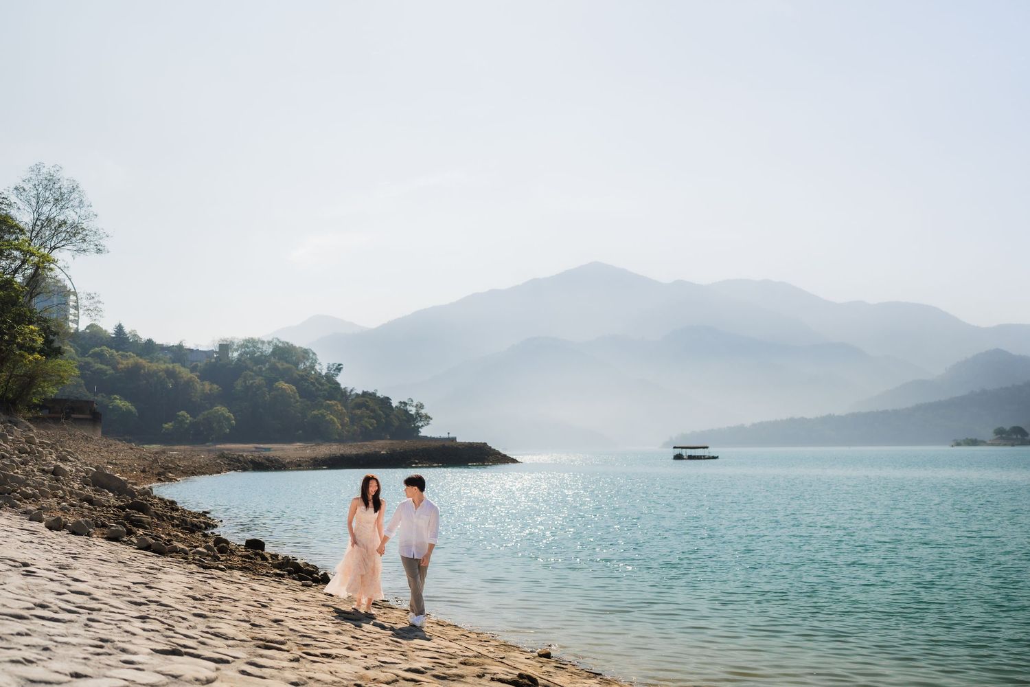 Couple walking hand in hand along a serene lakeside beach with misty mountains in the background.