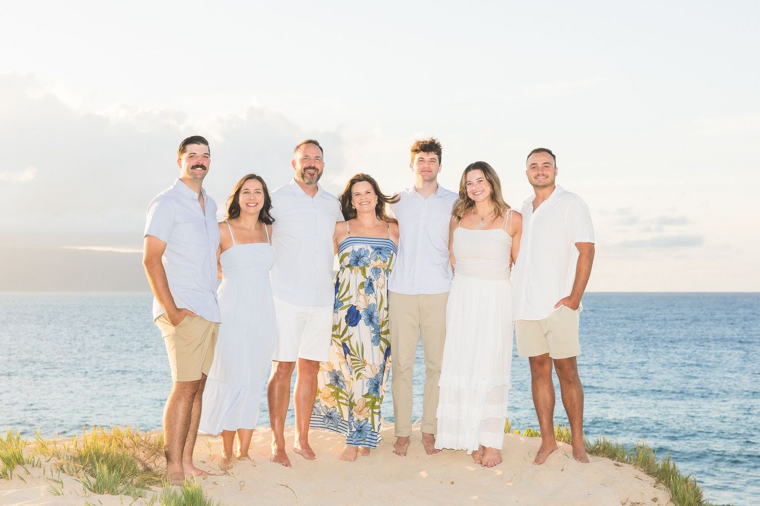 Large family group portrait on sandy beach dunes at sunset, wearing coordinated white and blue outfits.