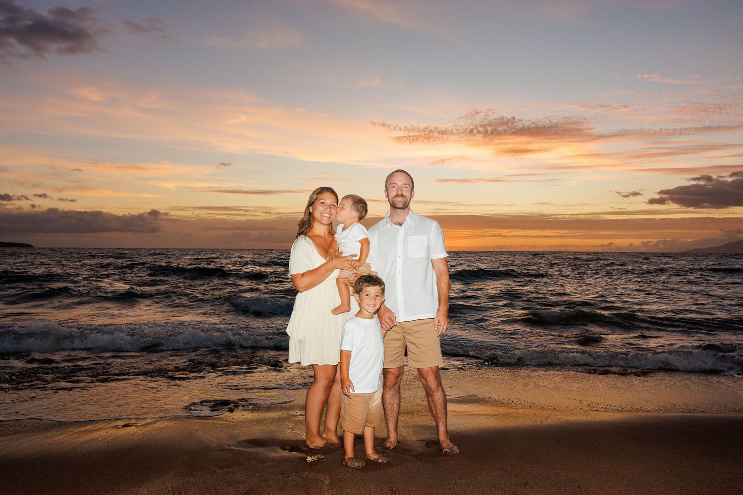 Family of four posing together on a beach during a stunning golden sunset over the ocean.