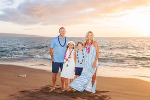 Family of four wearing leis posing on tropical beach at golden sunset in blue outfits.