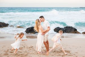 Family of four on a beach, parents embracing while two young children in white dresses play in the sand.