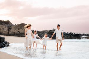 Happy family of four holding hands and walking through ocean waves on a tropical beach at sunset.