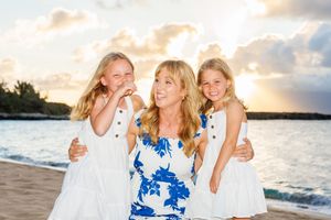 Smiling blonde mother posing with two young daughters in matching white dresses at sunset beach.