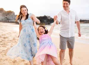 Laughing parents swinging happy little girl in colorful dress between them on sandy beach.