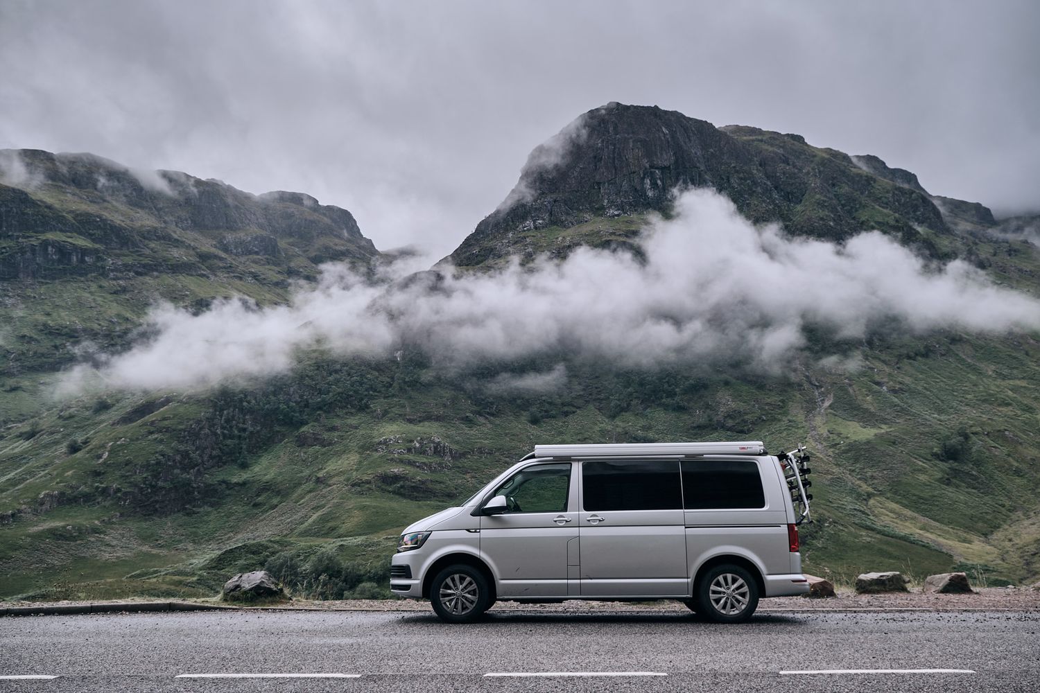 Silver Volkswagen Transporter campervan parked on road with dramatic cloudy mountain backdrop.