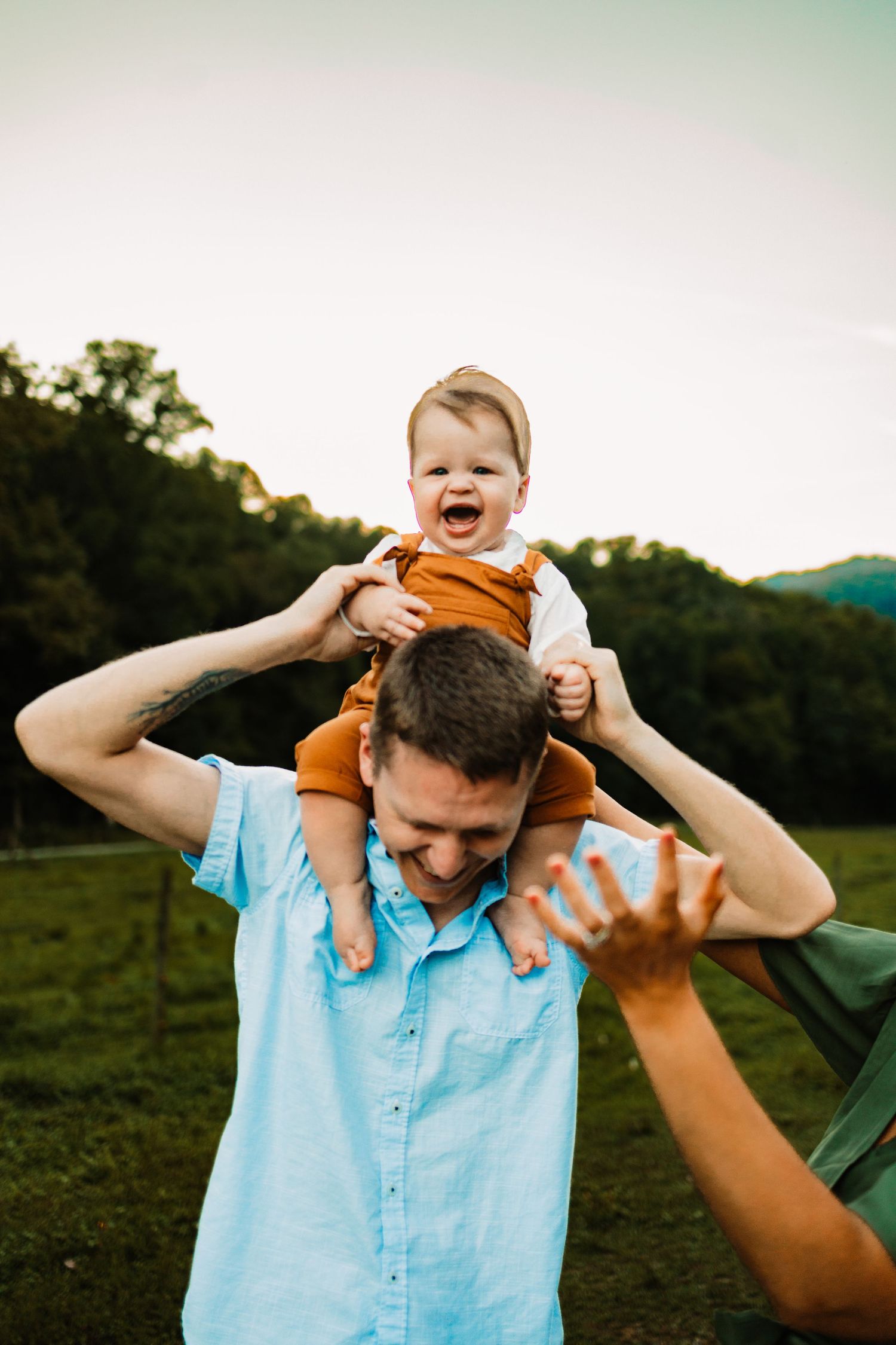 A laughing baby sits on a man's shoulders outdoors, surrounded by green hills and trees at dusk.