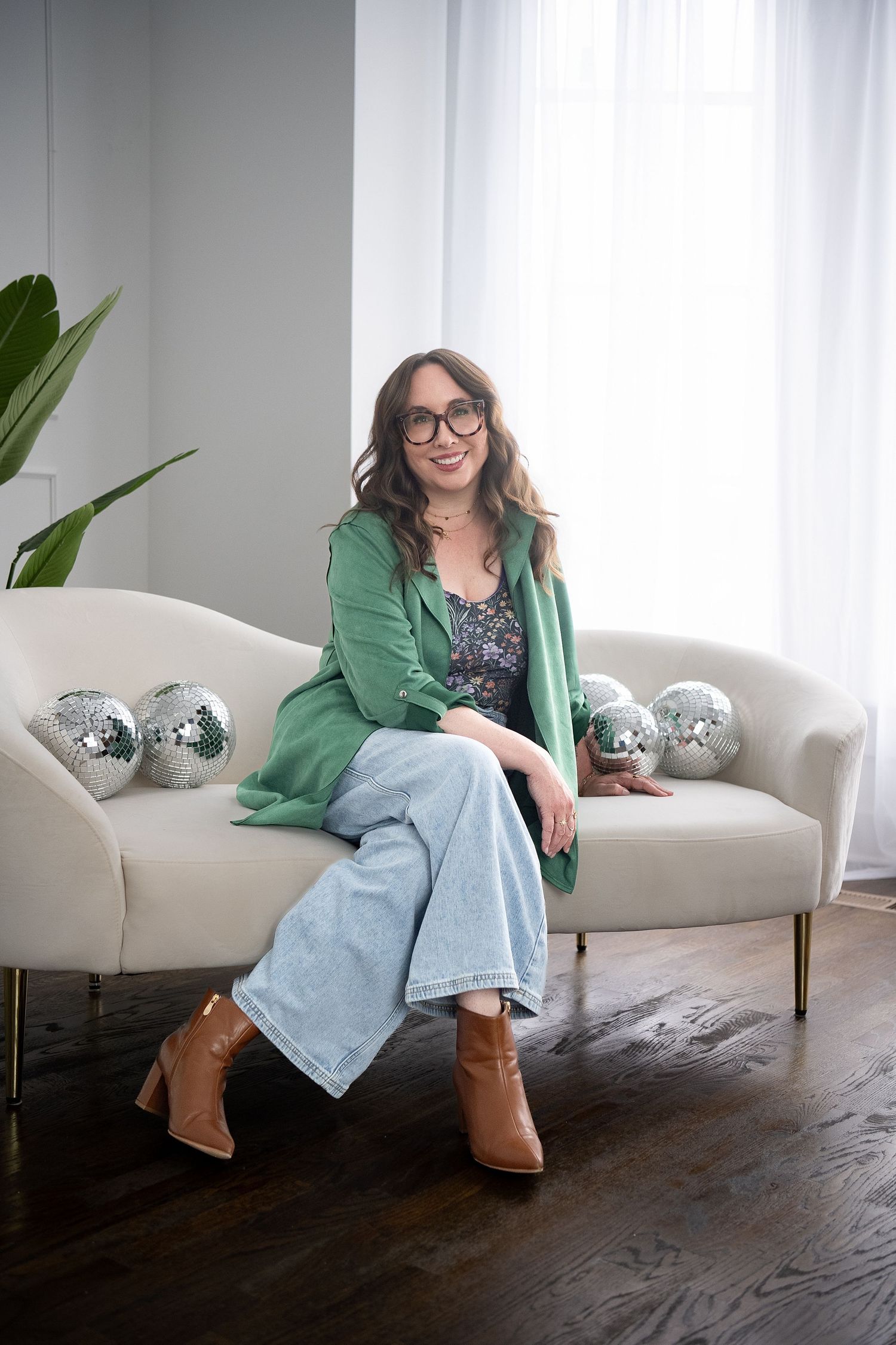 Smiling woman with glasses and wavy brown hair wearing a green blazer and floral top, arms crossed.