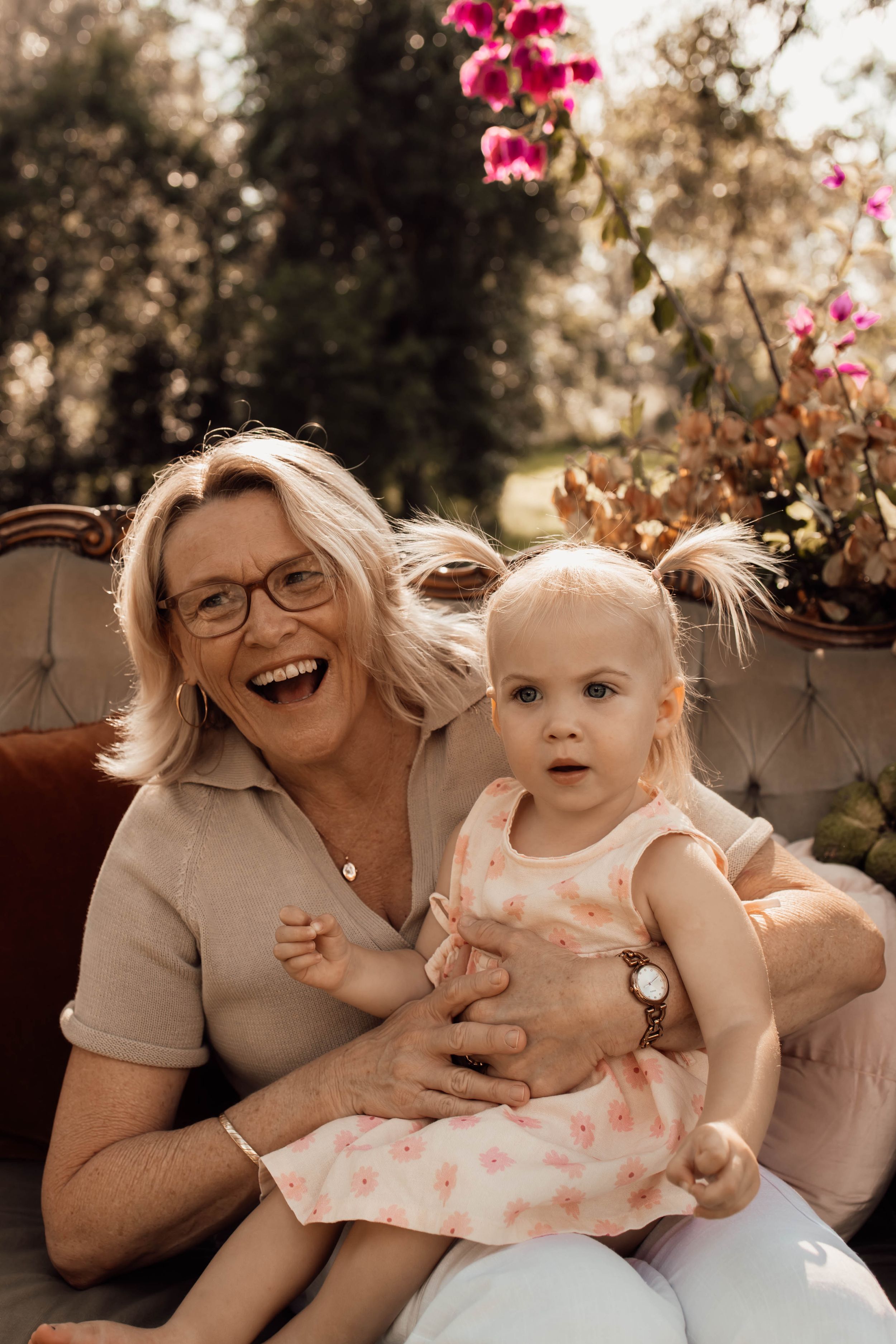Smiling grandmother with glasses holds a blonde toddler girl in a pink floral dress, seated outdoors near pink flowers.