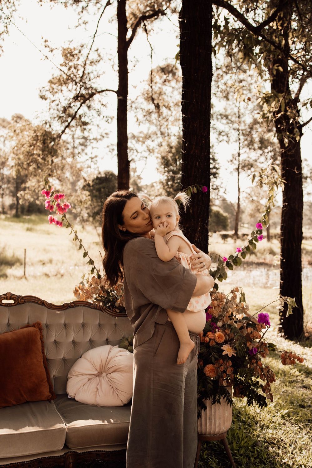 A woman in a gray dress holds a baby outdoors near a vintage sofa with colorful floral arrangements and trees.