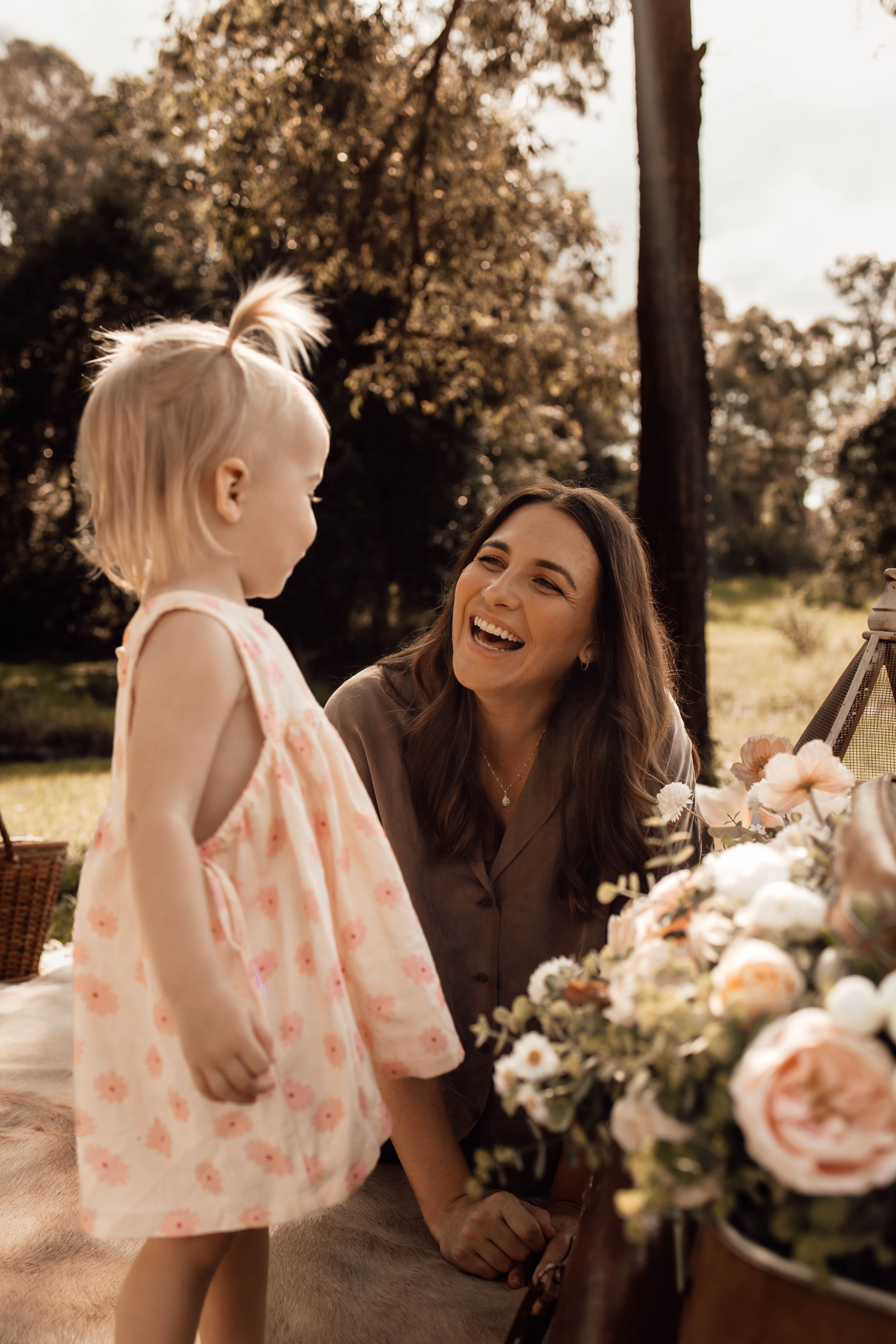 A laughing dark-haired woman crouches to interact with a toddler girl in a floral dress outdoors near flowers.