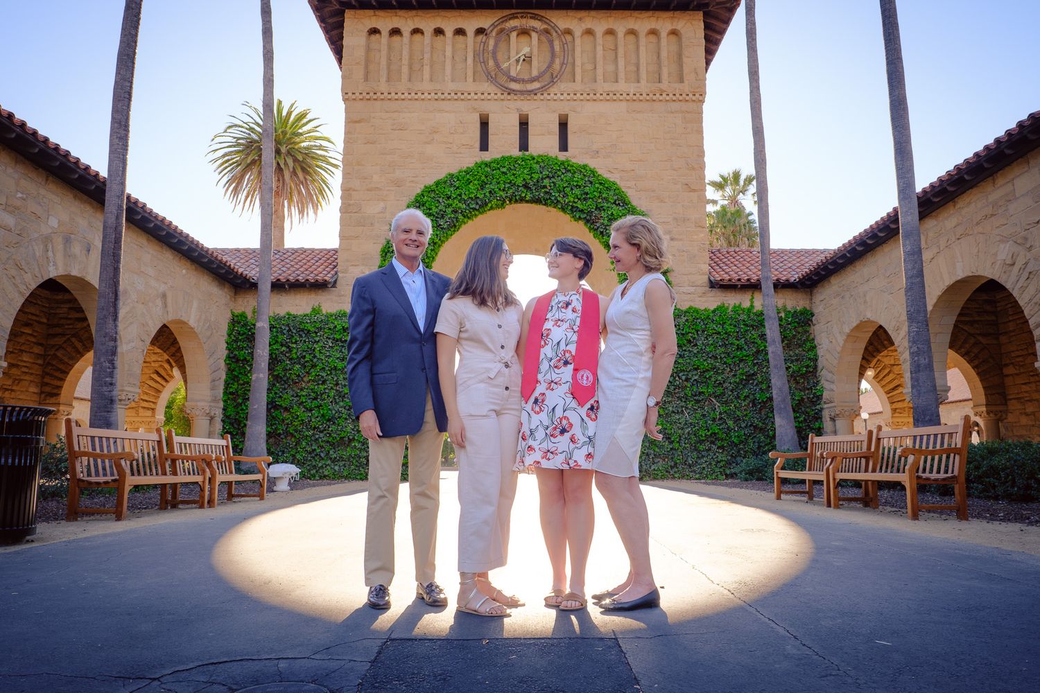Four people posing together in front of Stanford University's iconic arched sandstone building with lush ivy and palm trees.