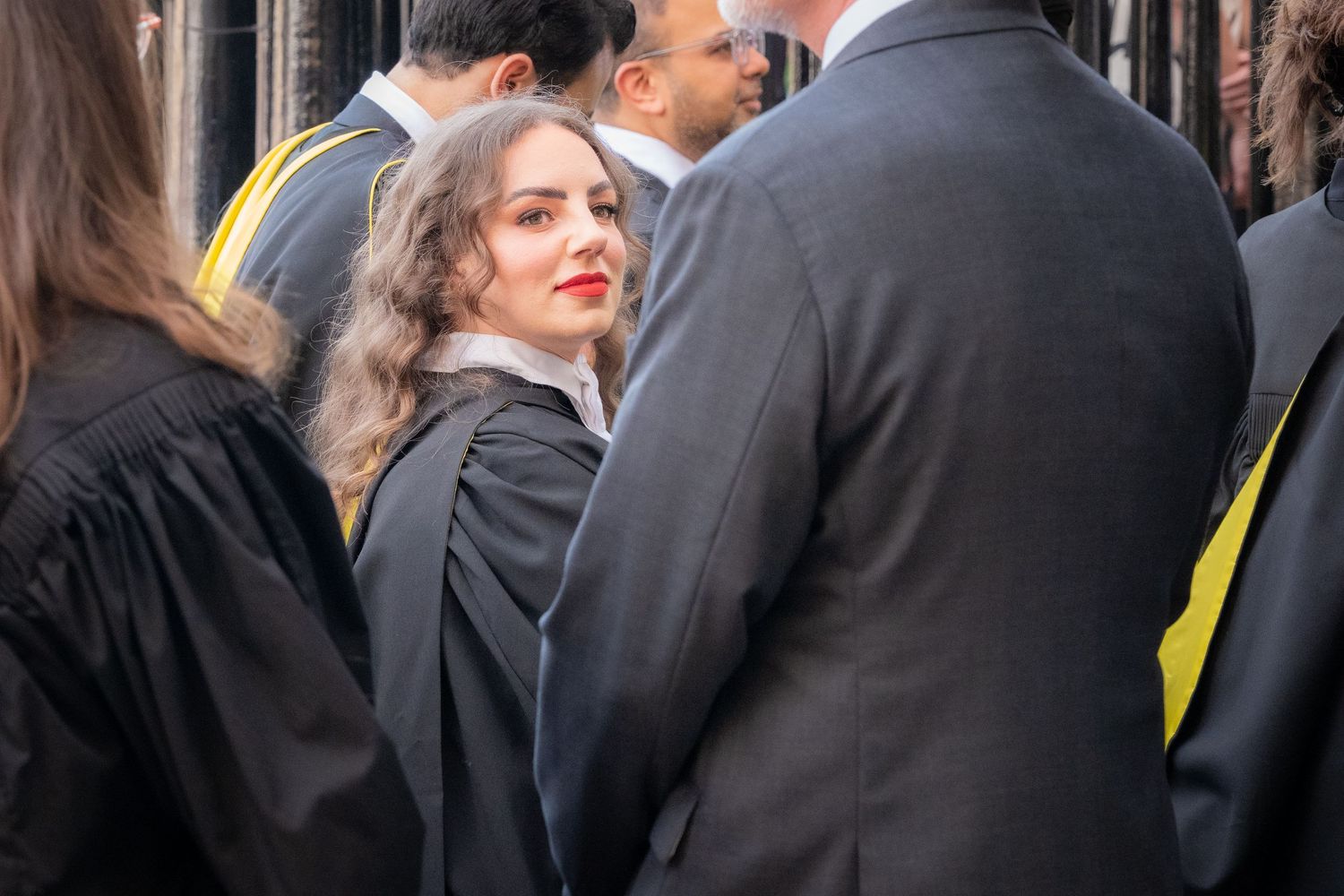 Young woman in graduation gown with red lipstick smiling among graduates at a university ceremony.