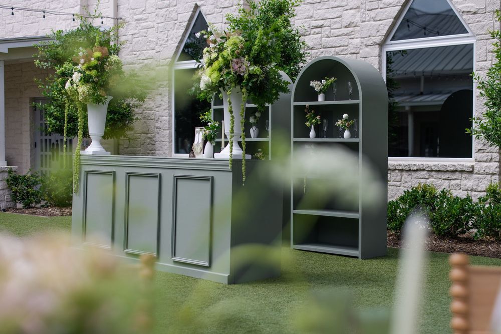 Elegant outdoor green bar setup with arched shelving unit, floral arrangements, and lush greenery at a garden wedding venue.