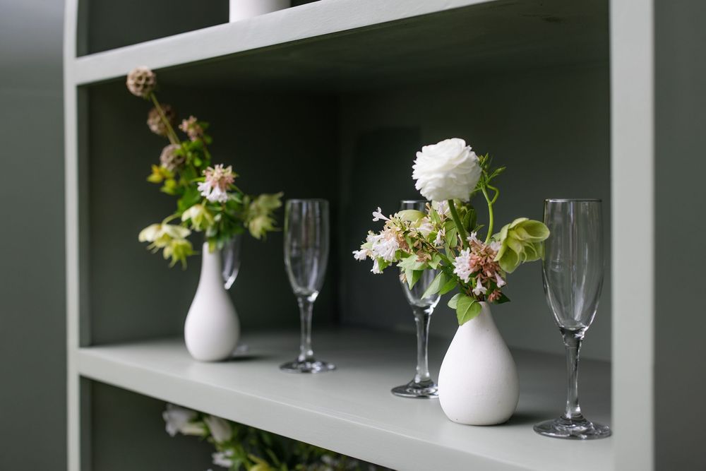 White vases with fresh flowers and champagne flutes displayed on sage green shelves for elegant home decor.