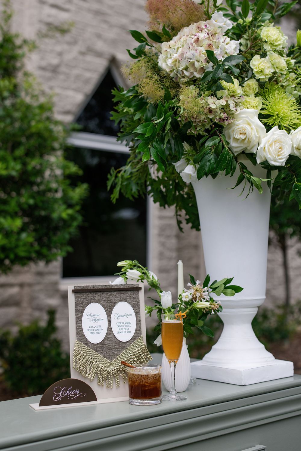 Elegant wedding bar display with white floral arrangement, signature cocktails, and decorative menu sign on sage green table.