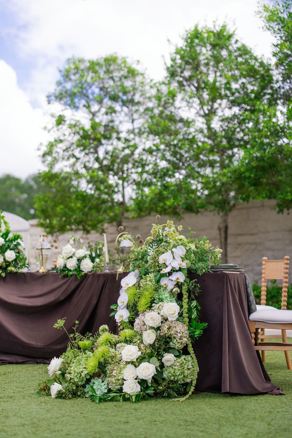 Outdoor wedding reception table with white roses and green floral arrangements on dark brown linen, set on grass.