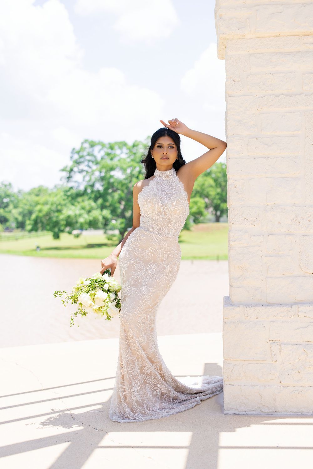 Bride in lace halter gown holding white bouquet leans against stone column outdoors near a pond.