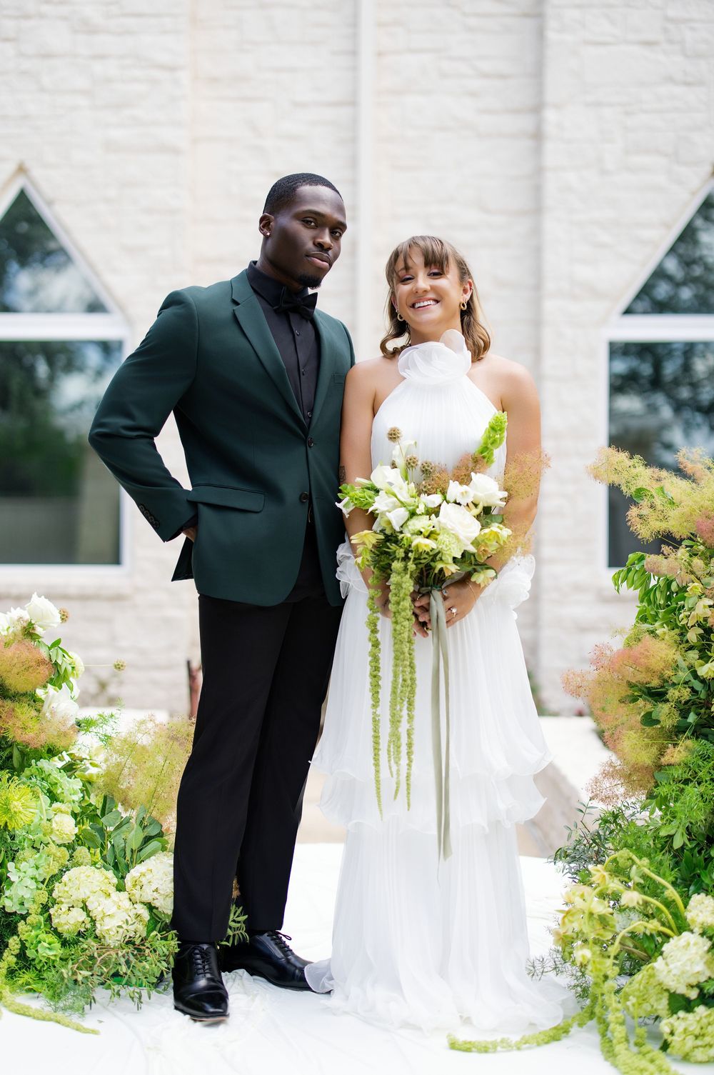 Interracial couple poses outdoors at church, groom in green suit, bride in white halter gown holding lush floral bouquet.