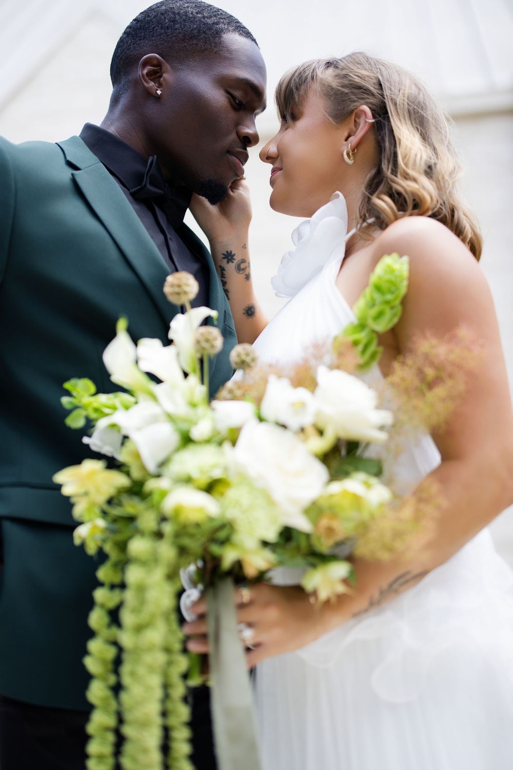 Interracial couple sharing romantic moment at wedding, groom in green suit, bride in white gown holding bouquet of white and green flowers.