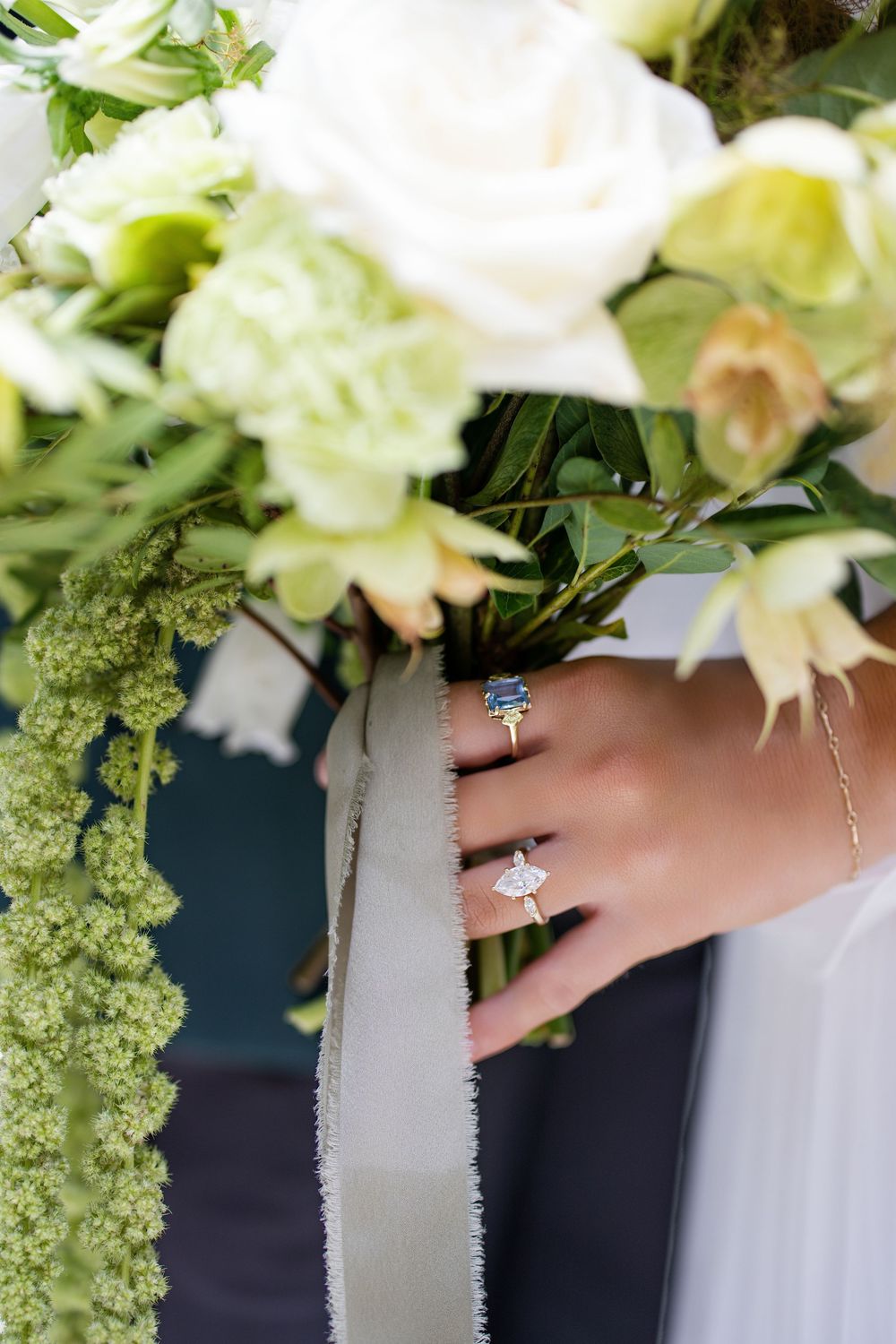 Bride holding white floral bouquet with ribbon, showcasing gold engagement ring and sapphire ring on wedding day.