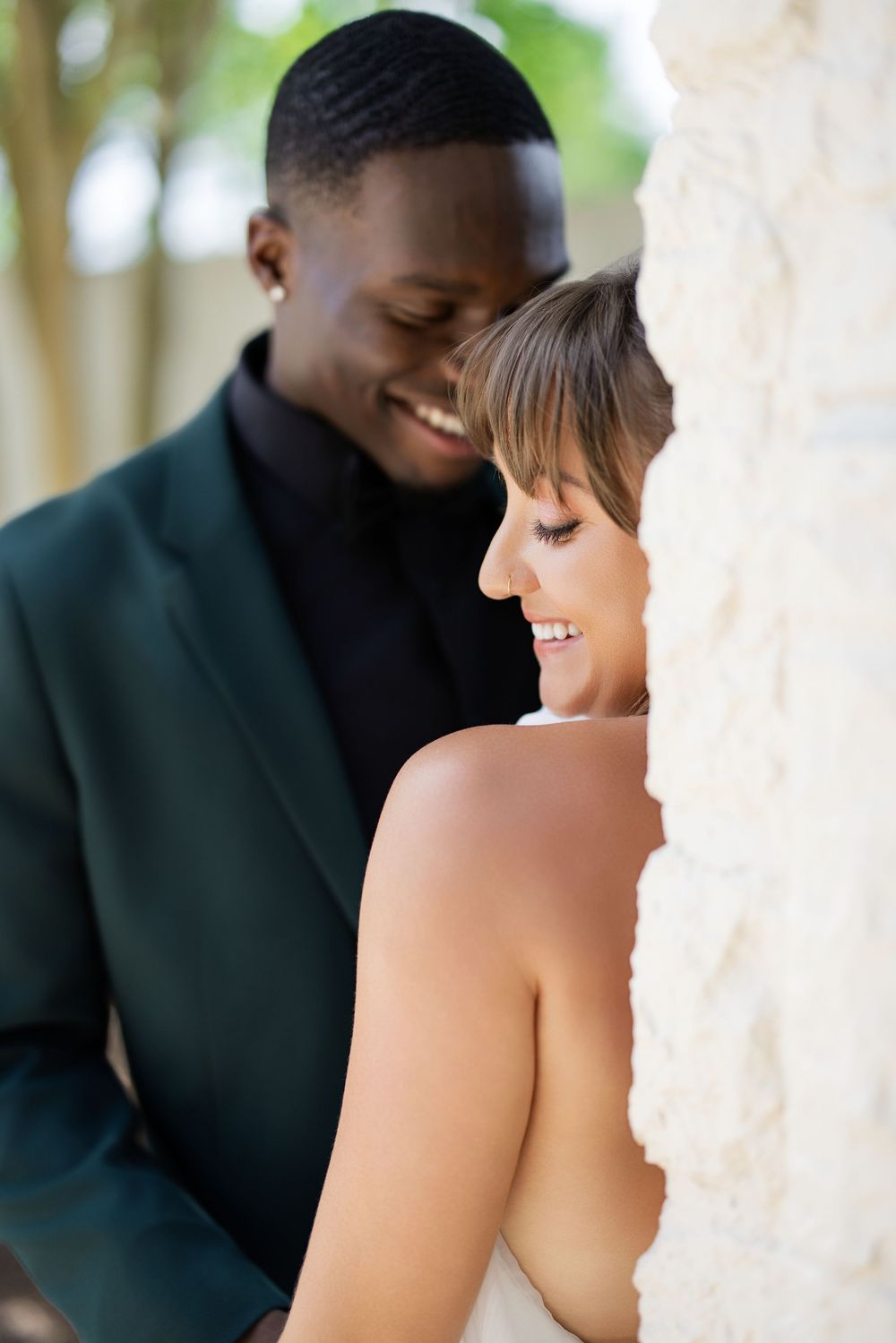 Smiling interracial couple in formal attire sharing a romantic moment near a stone pillar outdoors.