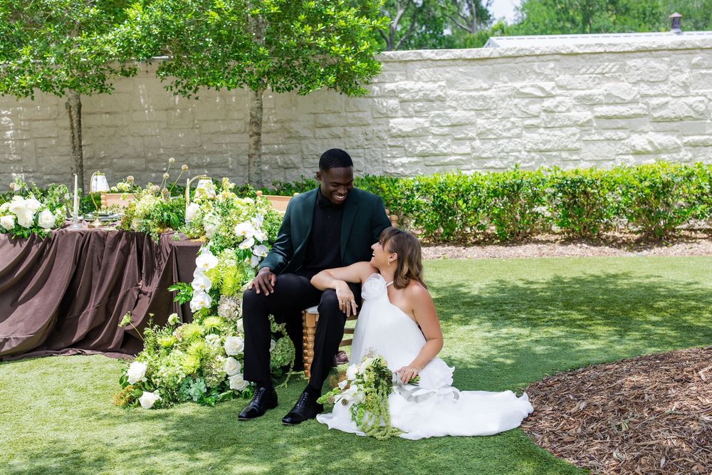 Bride in white gown sits on grass beside groom in black suit during outdoor wedding ceremony with floral arrangements.