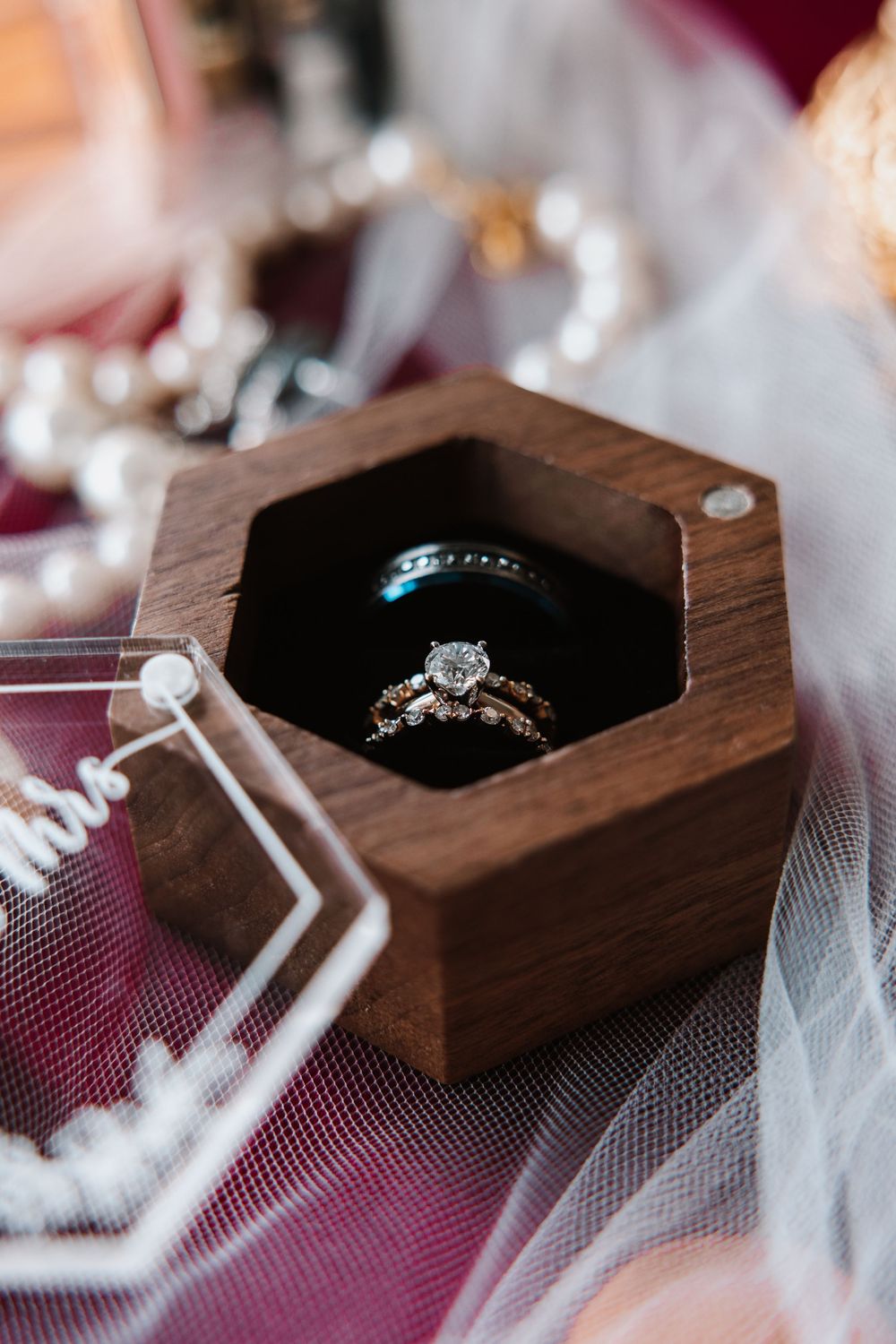 Diamond engagement ring and wedding bands displayed in a hexagonal wooden ring box on a burgundy velvet surface.