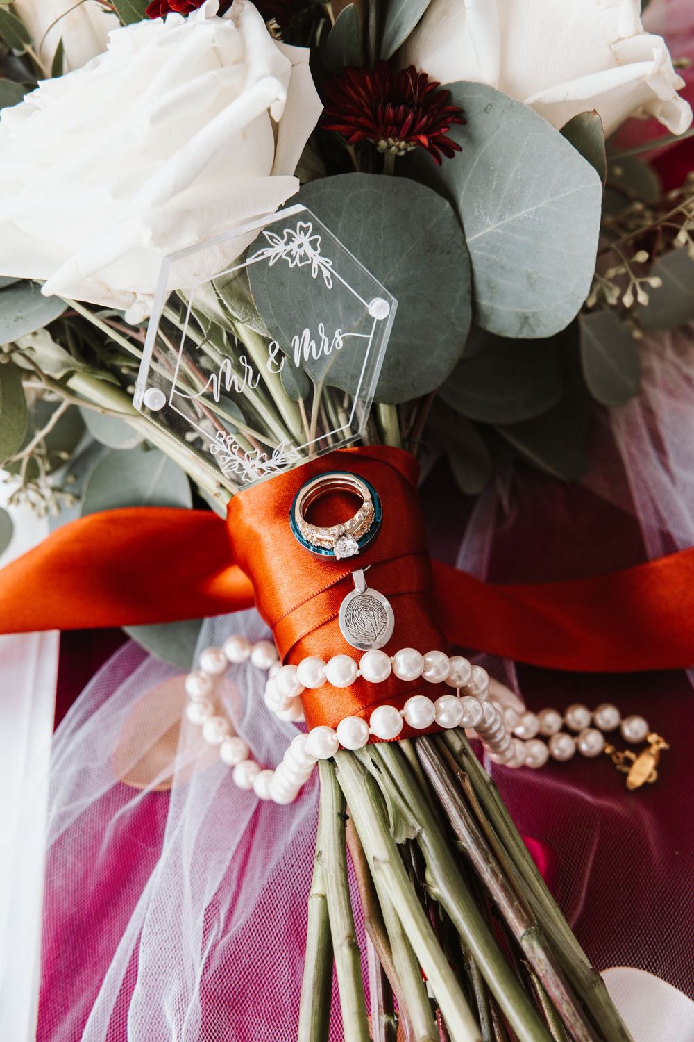 Bridal bouquet with white roses, eucalyptus, pearl bracelet, wedding rings, and Mr. & Mrs. acrylic sign tied with orange ribbon.