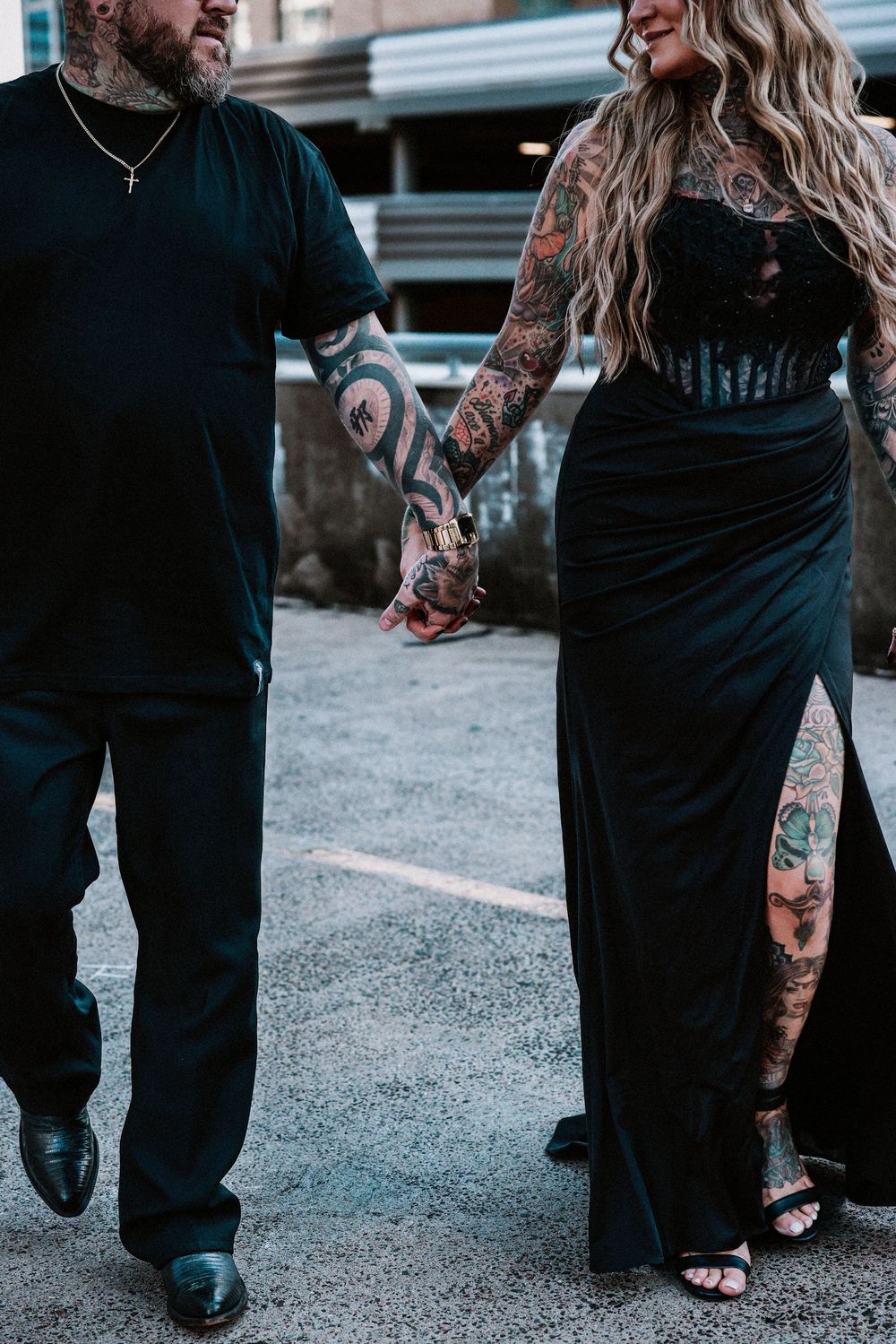 Tattooed couple holding hands in black formal attire, walking outdoors on a paved surface.