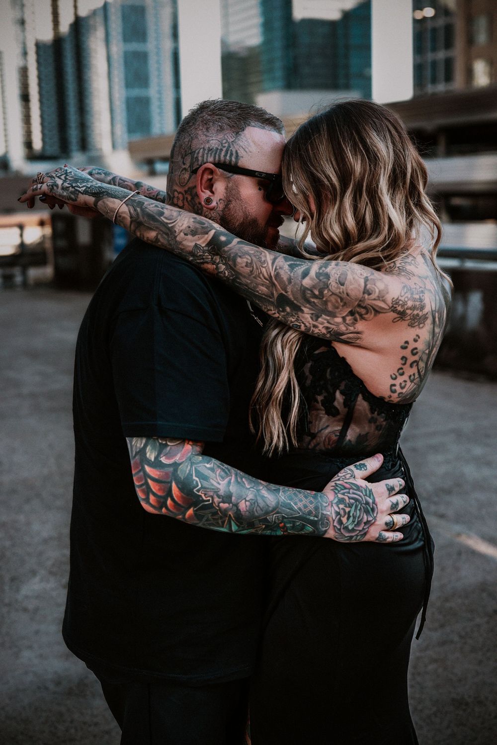 Heavily tattooed couple embracing and kissing outdoors in an urban setting, both wearing black clothing.