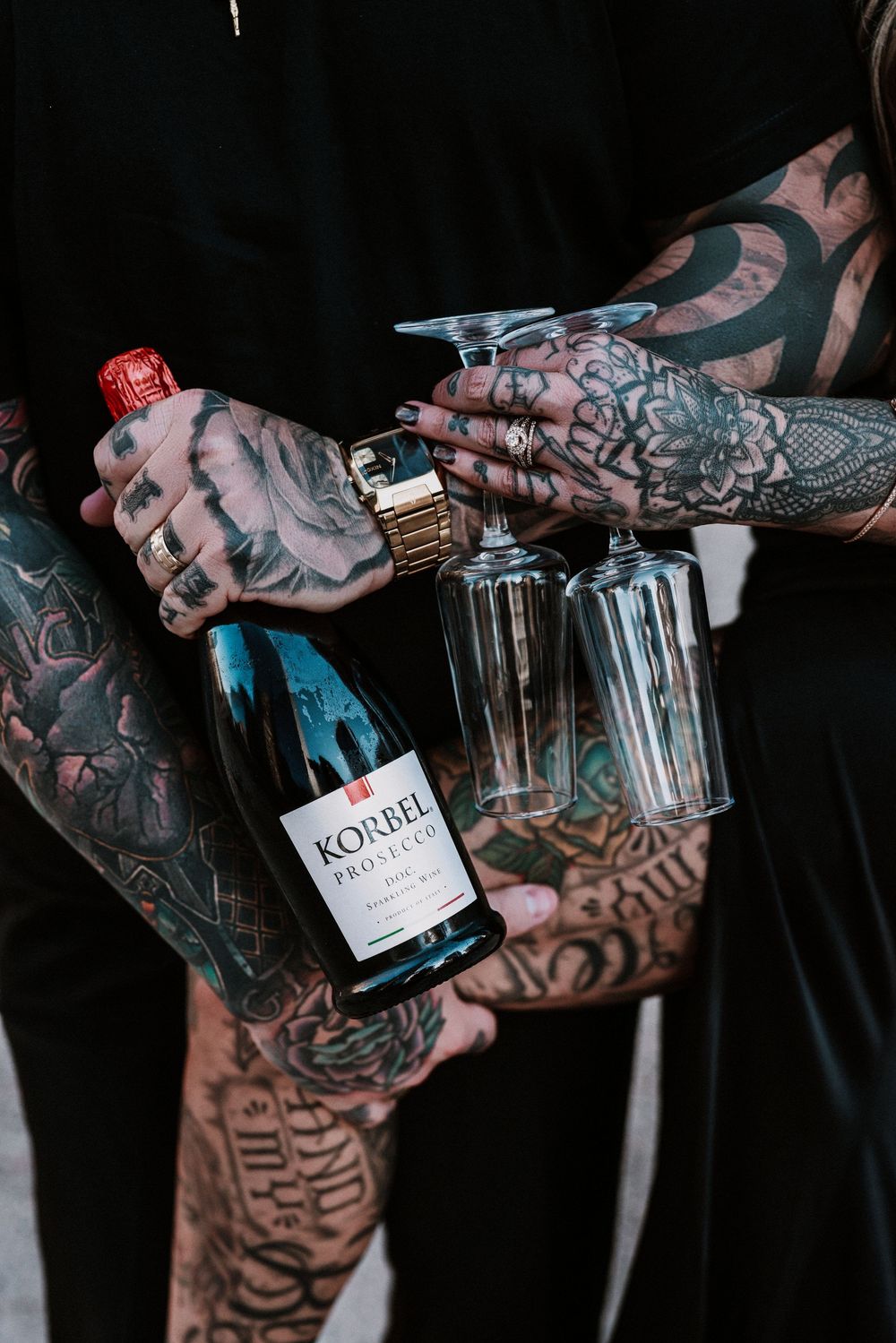 Heavily tattooed hands holding a Korbel Prosecco bottle and two champagne flutes against a dark background.