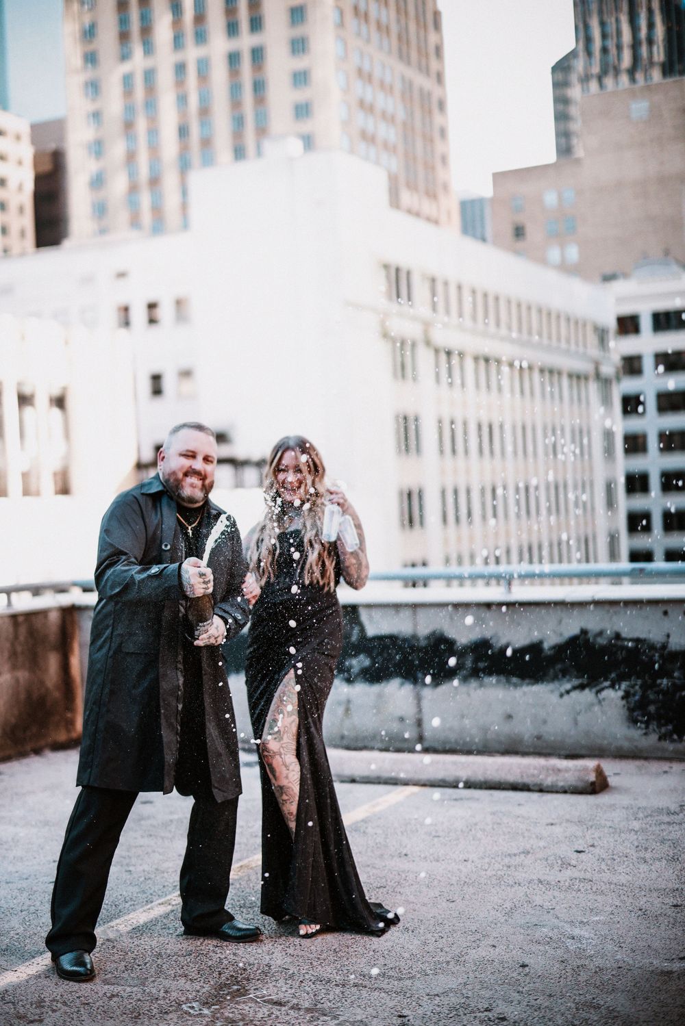 Couple in black formal wear celebrating on a rooftop with confetti, city buildings in the background.