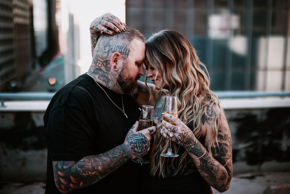 Tattooed couple sharing a romantic moment clinking champagne glasses on a rooftop, dressed in casual black attire.
