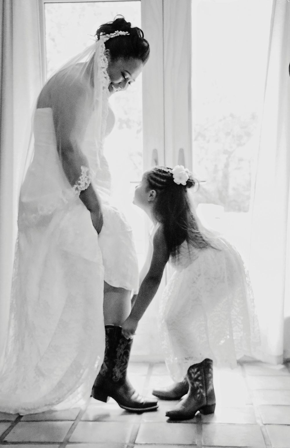 Black and white photo of a bride in cowboy boots with a flower girl in matching boots helping her prepare.
