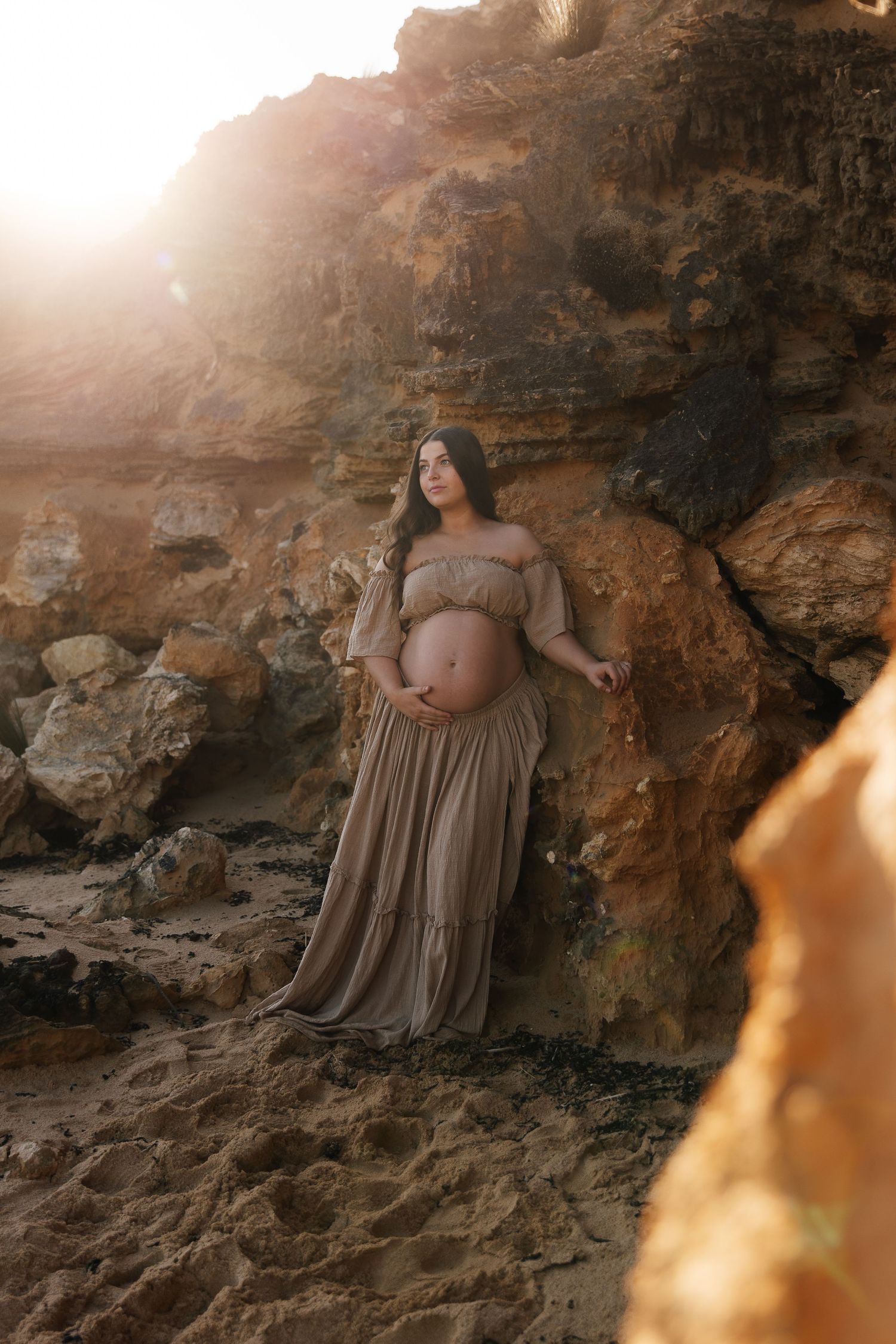 Pregnant woman in flowing tan off-shoulder gown poses against golden rocky cliffs during sunset maternity photoshoot.