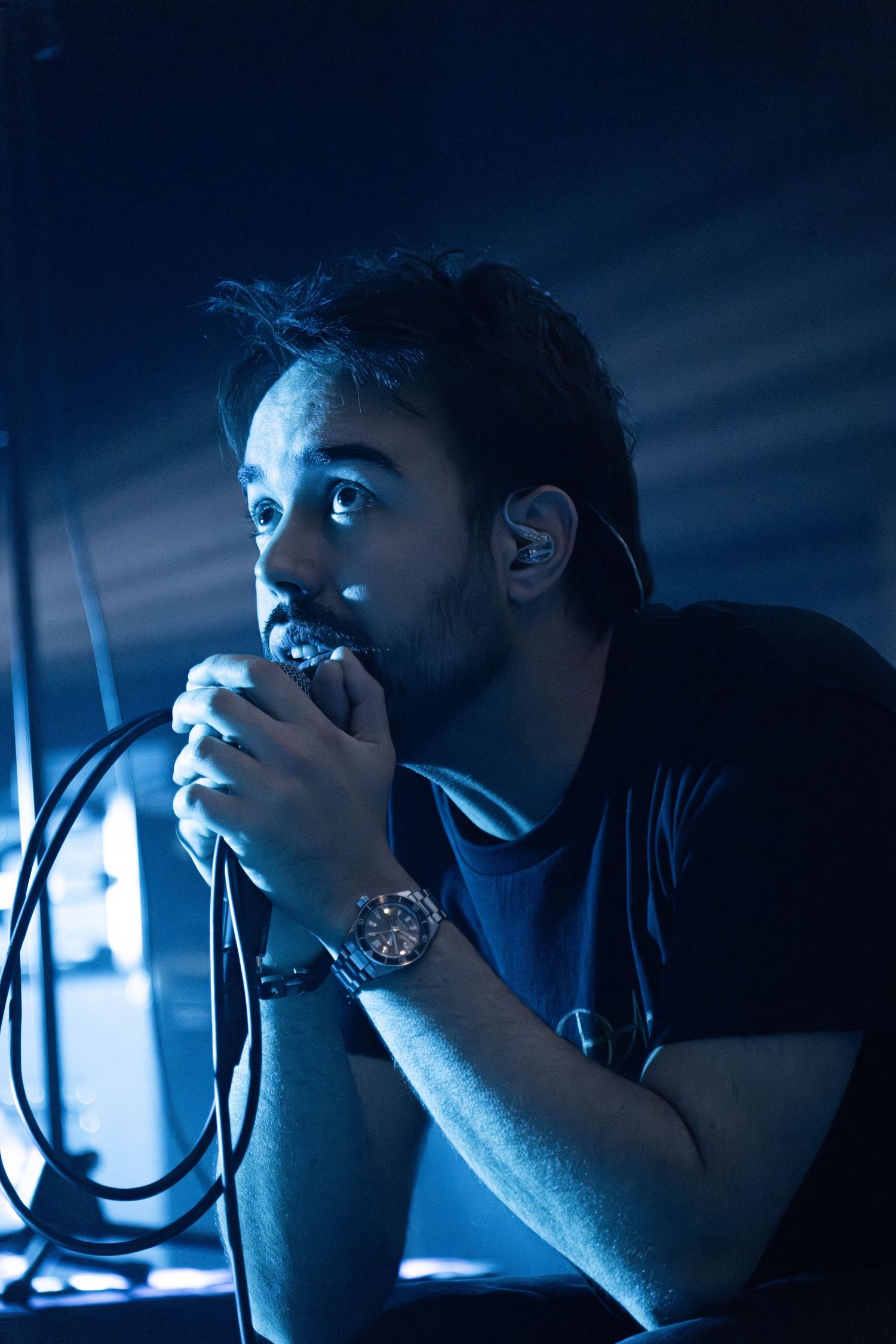 Male singer performing on stage under blue lighting, holding microphone close to mouth, wearing a watch and in-ear monitor.