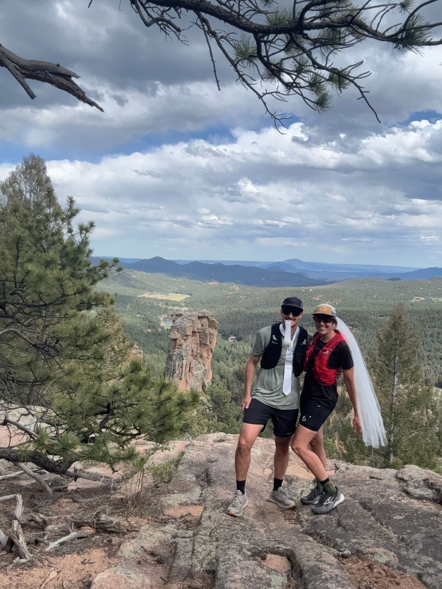 Couple in hiking gear with bridal veil posing on rocky overlook with panoramic mountain forest views under cloudy sky.
