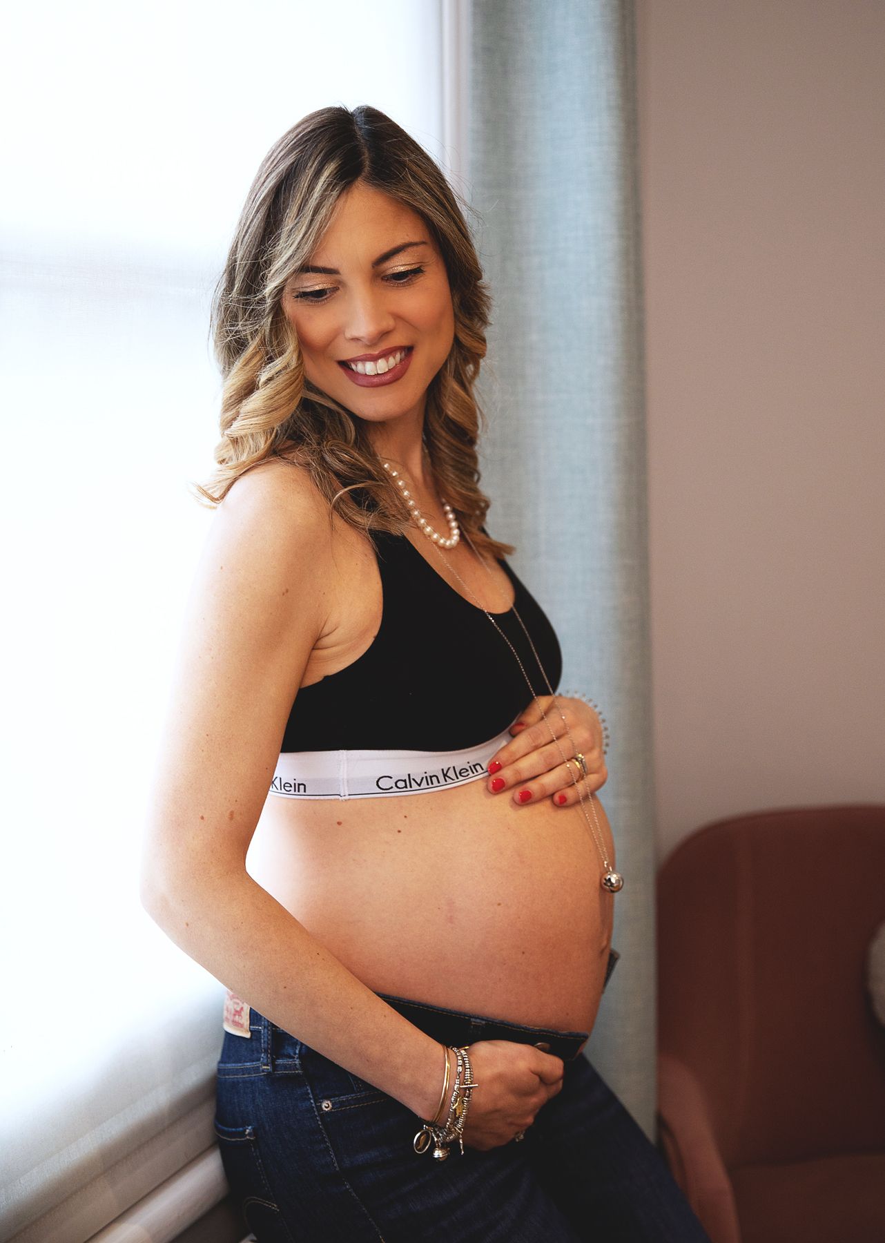 Smiling pregnant lady in black Calvin Klein sports bra and jeans cradling baby bump, wearing pearl necklace. During a home maternity session