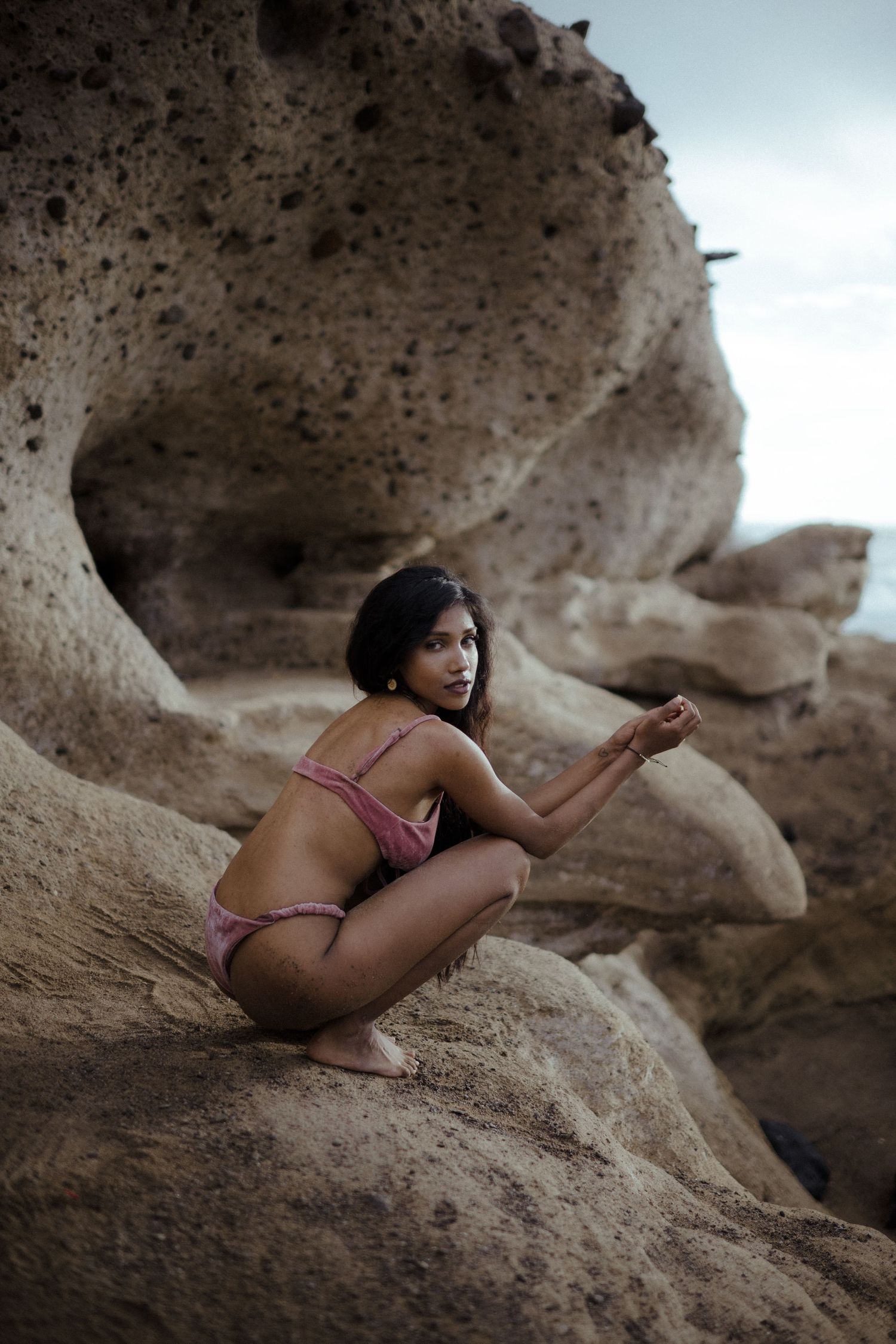 Woman in pink bikini posing gracefully on rocky coastal formations with dramatic stone textures in background.