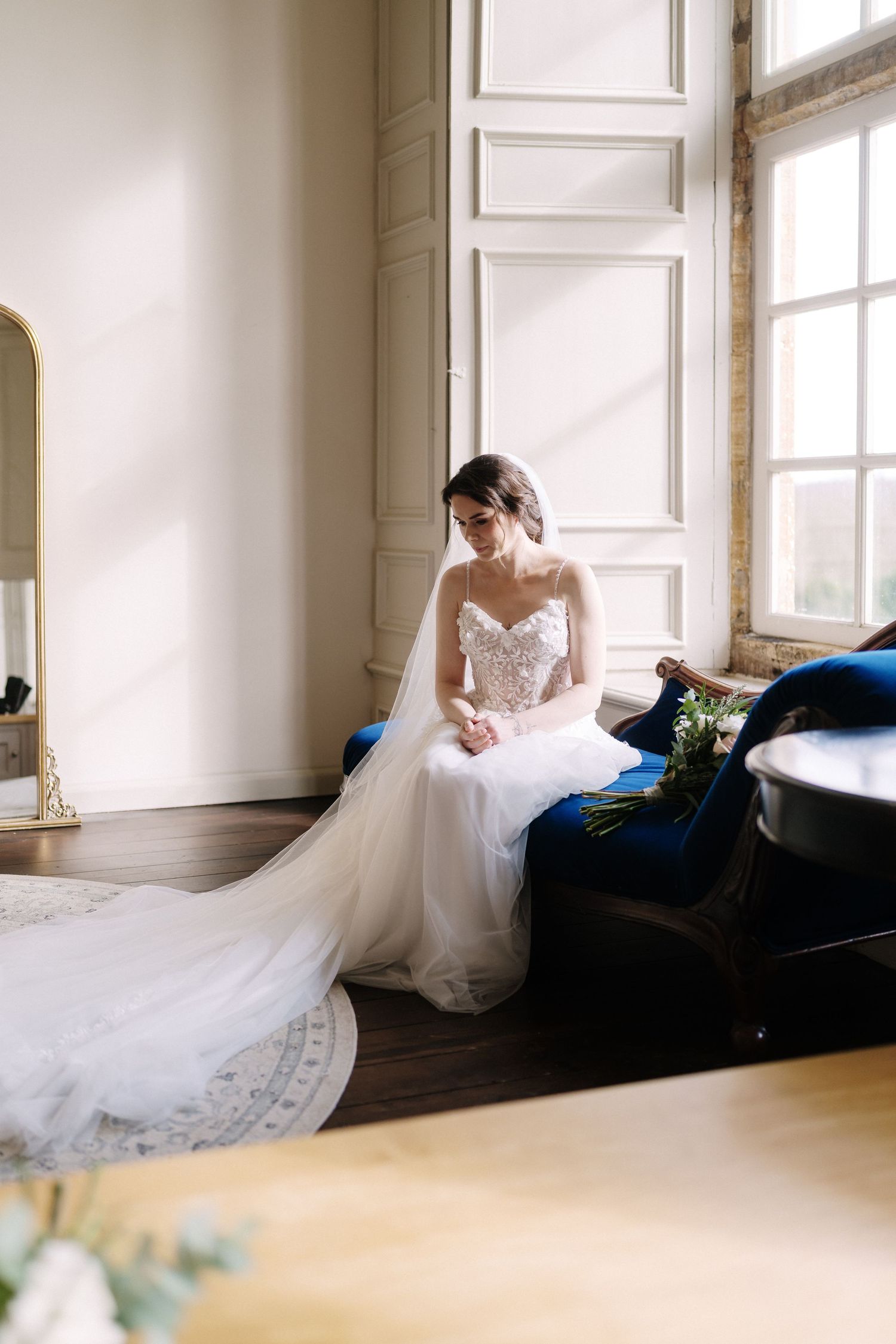 Bride in a beaded strapless gown with long veil sits on a blue velvet chair by a sunlit window holding a bouquet.