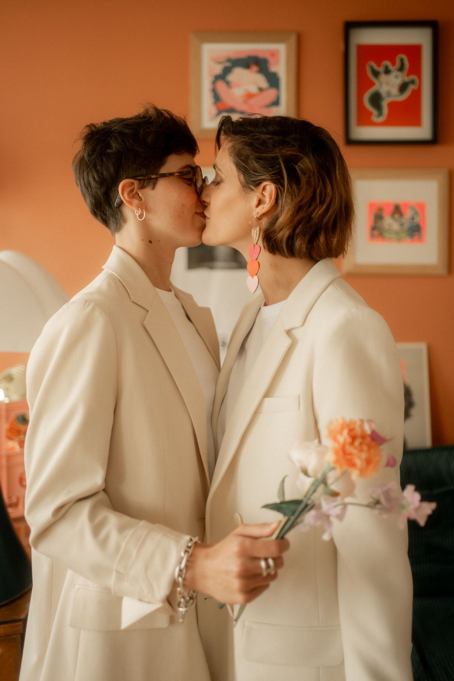 Two women in white blazers sharing a kiss, one holding flowers, in a warm room with colorful art prints.